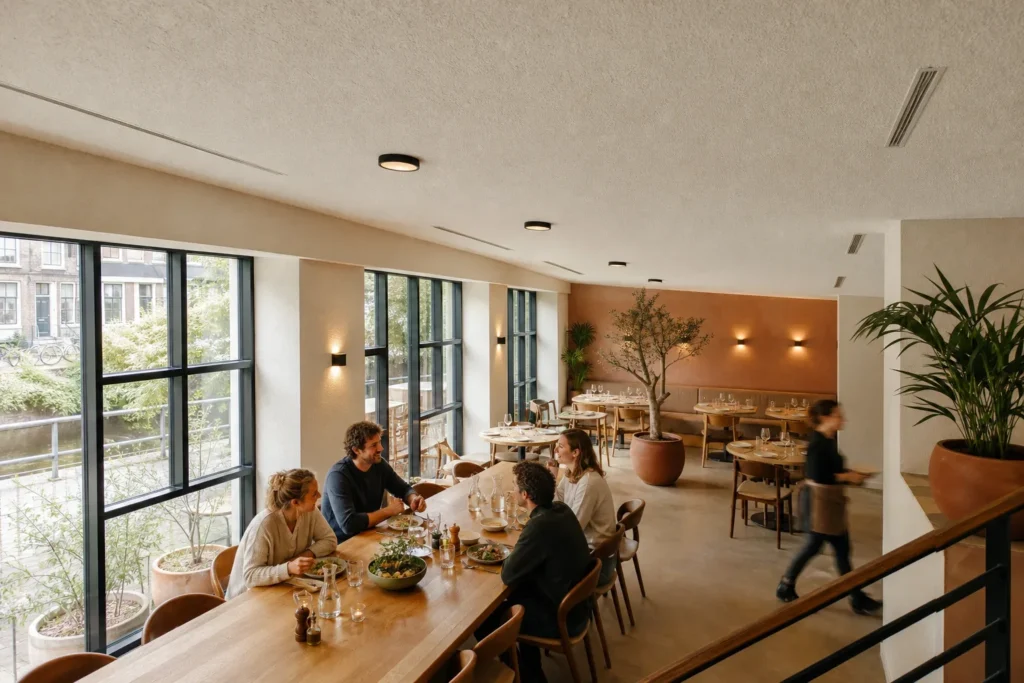 Van analyse naar een passende restaurantoplossing: Vier volwassen gasten zitten verspreid aan de lange tafel in natuurlijk gesprek; een bedieningsmedewerker beweegt zacht onscherp langs de rand van het beeld. Geen logo’s, merknamen; De gast